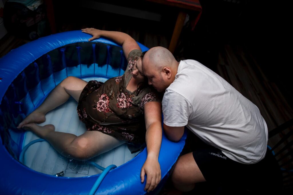 An expectant mother birthing in an inflatable birthing pool. Her partner sits near providing support.