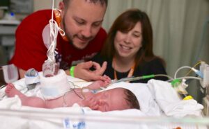 A newborn surrounded by medical equipment while the parents look on happily. Baby's hand is wrapped around a parent's finger.