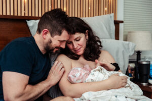 A new mother cradles her baby against her chest while the other parent looks over her shoulder.