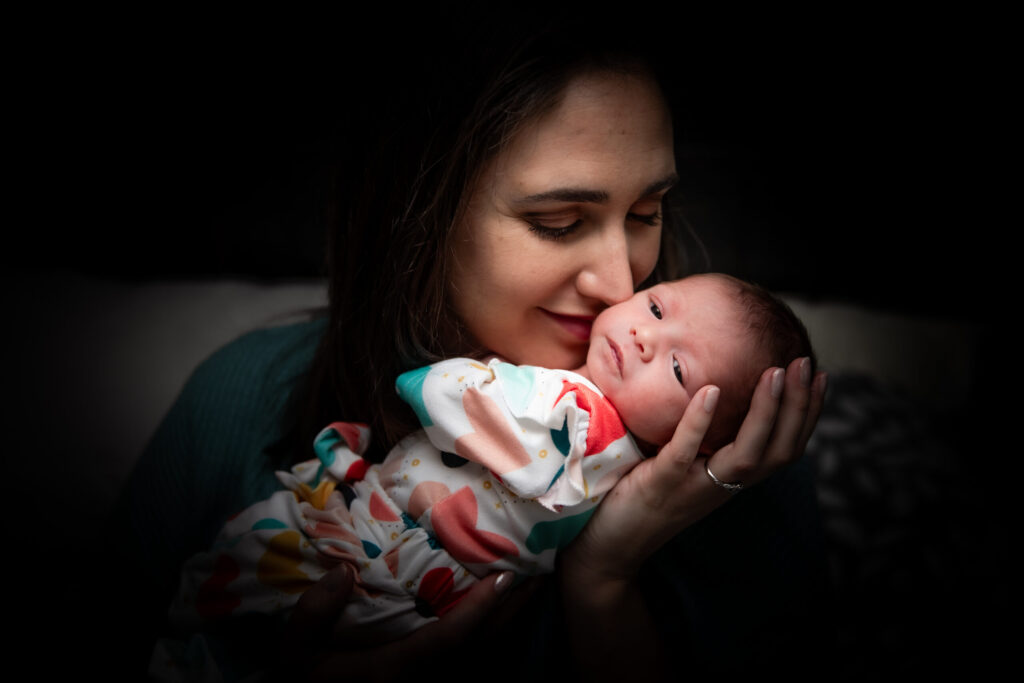 Woman holding a sleeping baby wrapped in a colorful blanket against a dark background.