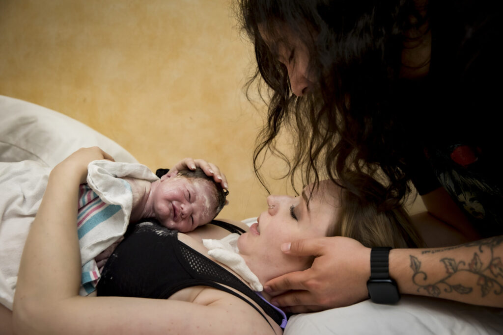 Woman holding and looking tenderly at a newborn baby in her arms while her doula gently massages her scalp to help her relax after giving birth