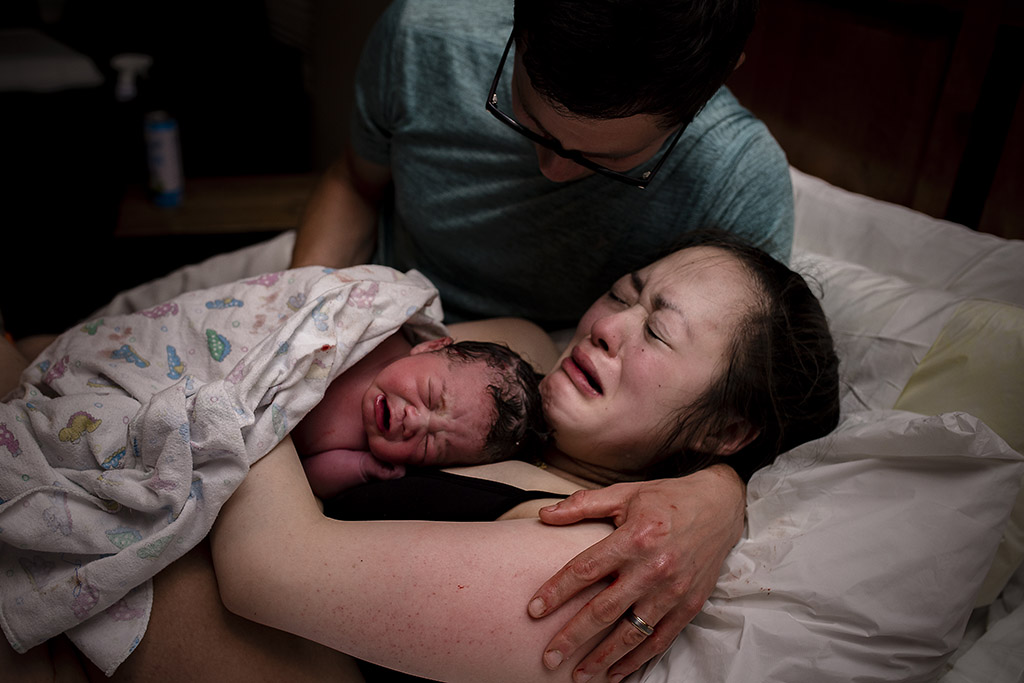 A woman experiencing post-birth emotions lying in a hospital bed, holding her newborn baby wrapped in a blanket, with a supportive partner beside her.