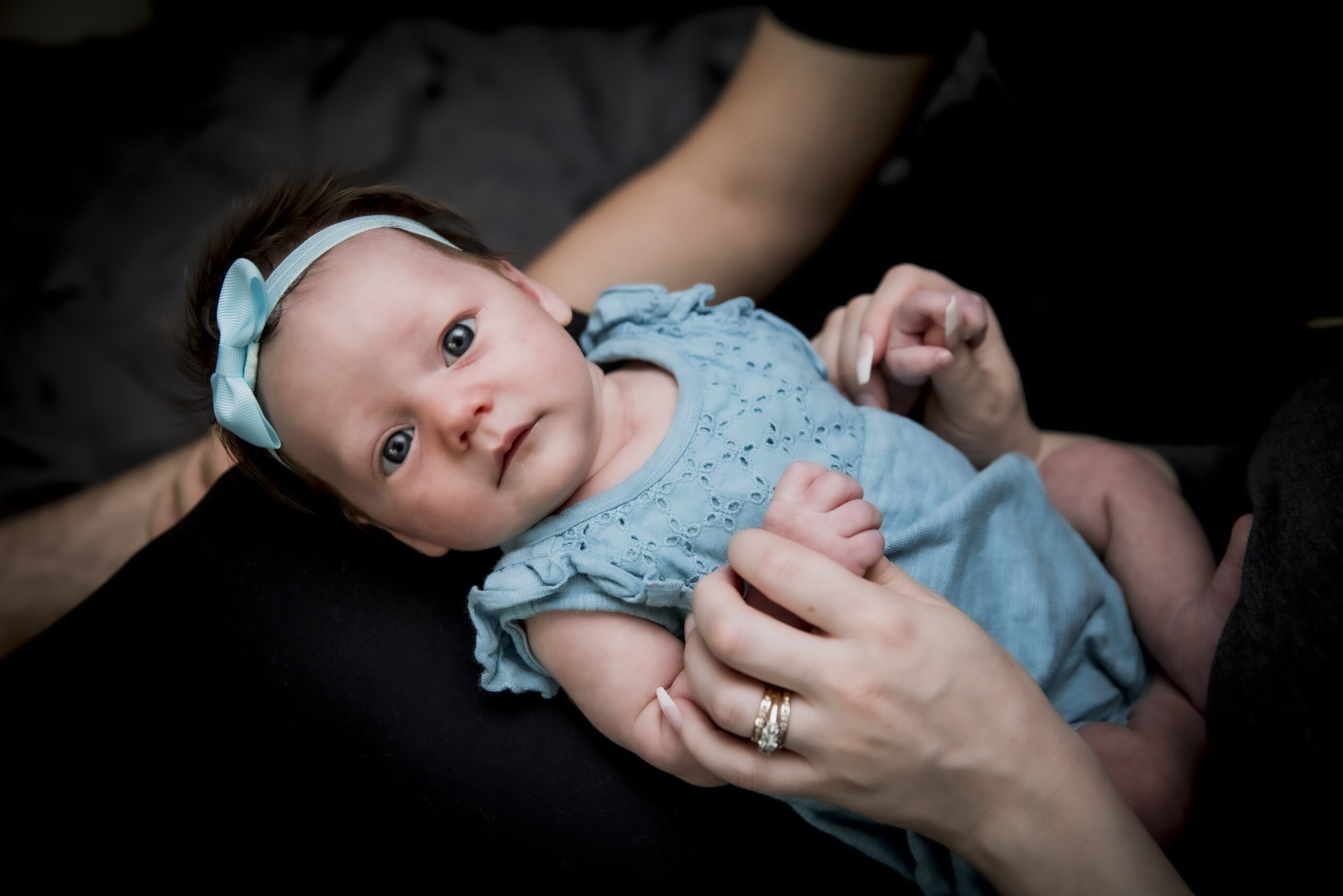 A baby in a blue outfit held securely in an adult's arms, against a black background.