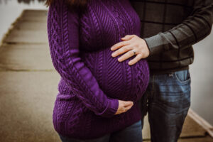 A pregnant person in a purple sweater with partner's hand embracing the belly from behind.