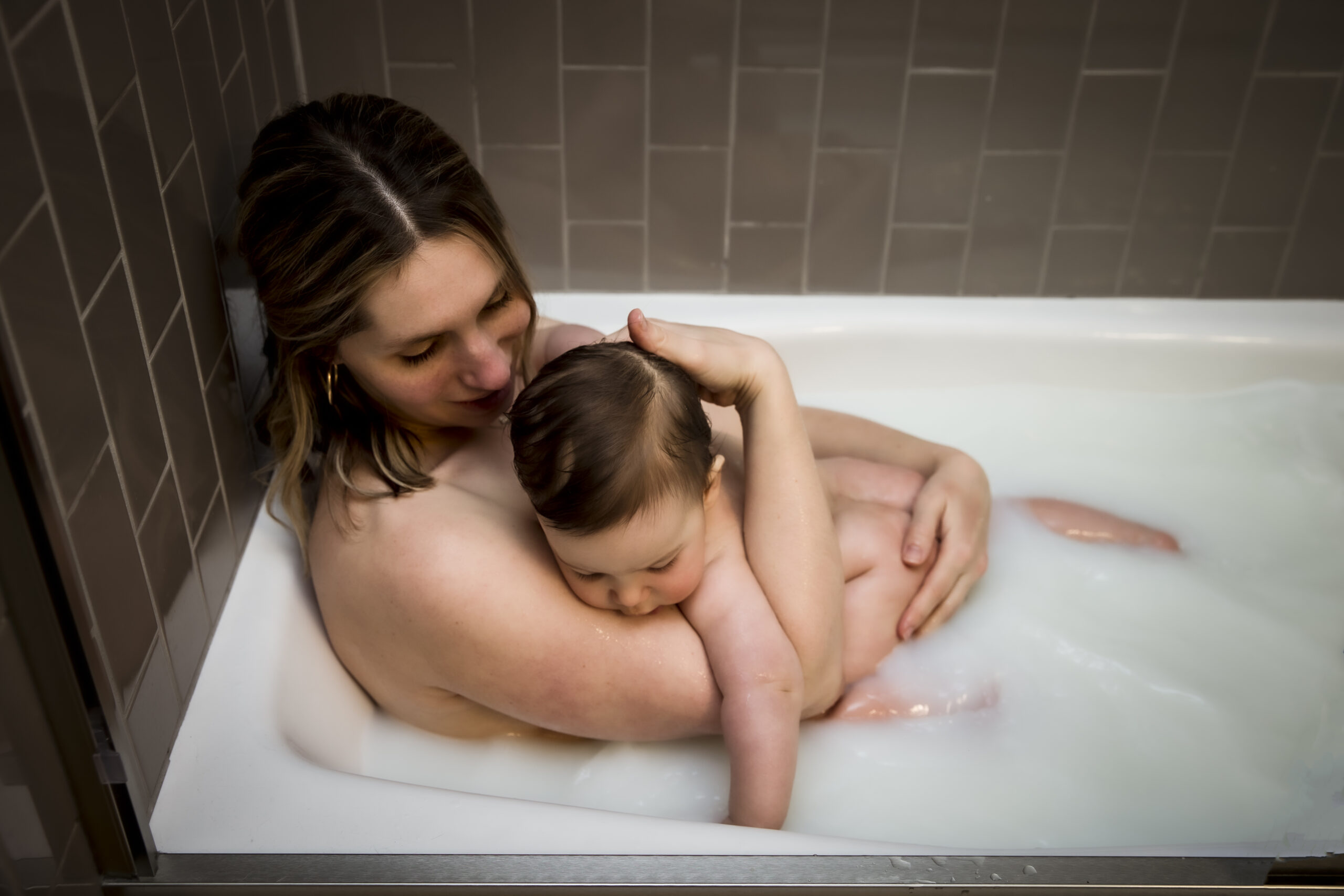 A woman embracing a child in a bathtub for a milkbath photograph, with brown tiled wall behind.