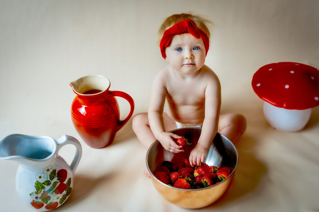 Child sitting with a bowl of strawberries, flanked by a red pitcher and toy mushroom.