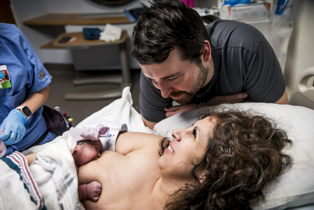 A newborn lies on a mother's chest nursing in a hospital room with the father standing over her shoulder