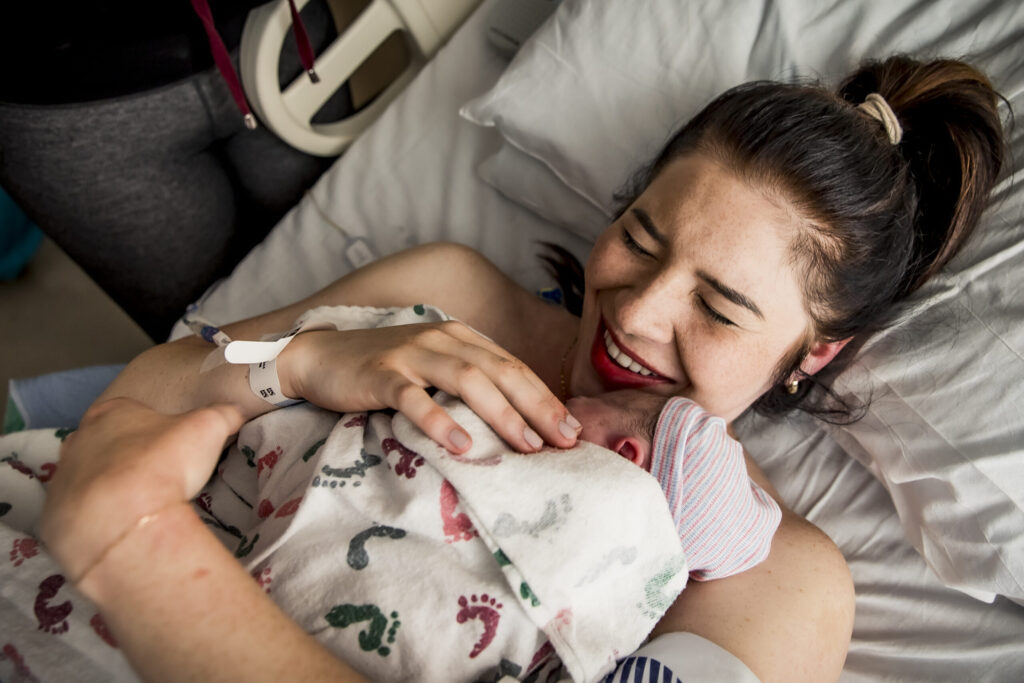 A new mother joyfully holds her newborn baby wrapped in a white hospital blanket, lying on a bed in a hospital room. The mother, wearing a hospital wristband, smiles with eyes closed, illustrating a tender moment of bonding and happiness. Keywords: new mother, newborn baby, hospital blanket, hospital room, joyful bonding, tender moment, happiness.