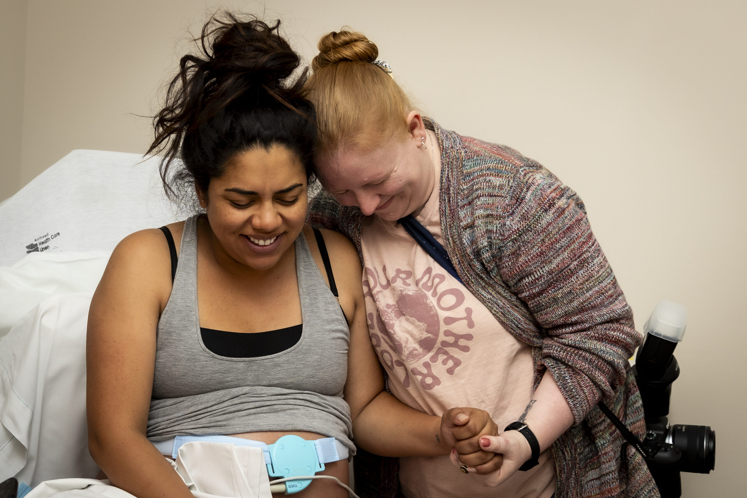 A birthing parent in a hospital bed wearing a monitor, her birth support doula is holding her hand and encouraging her through her birth