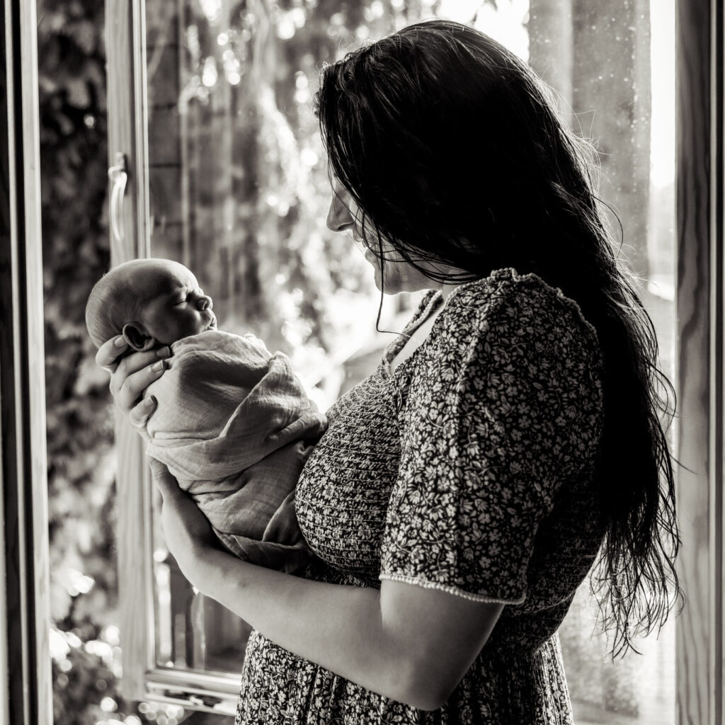 Woman in a floral dress holding a baby, standing by a window with trees outside.