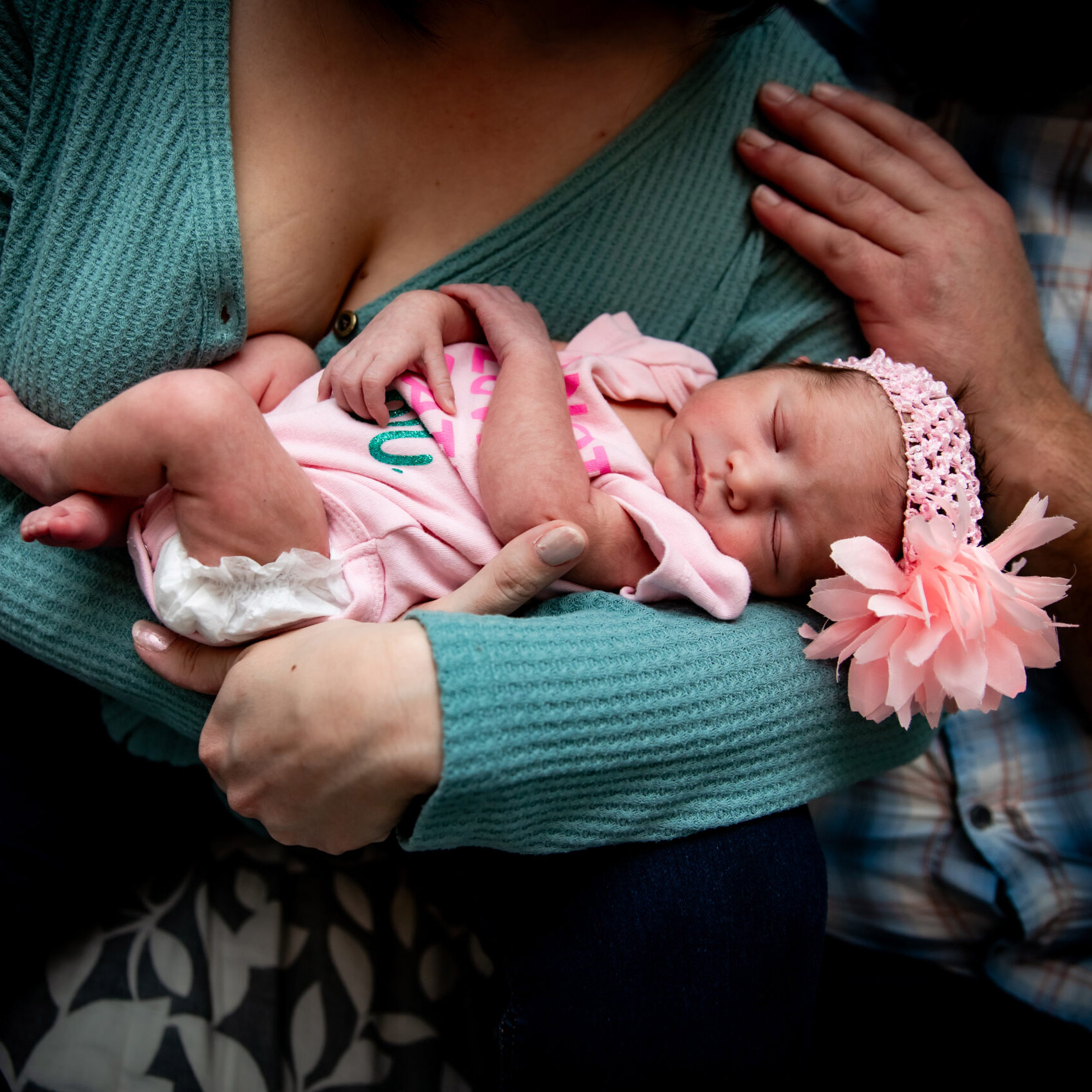 A baby in a pink outfit cradled by a woman in a teal sweater, with a man's hand on her shoulder.