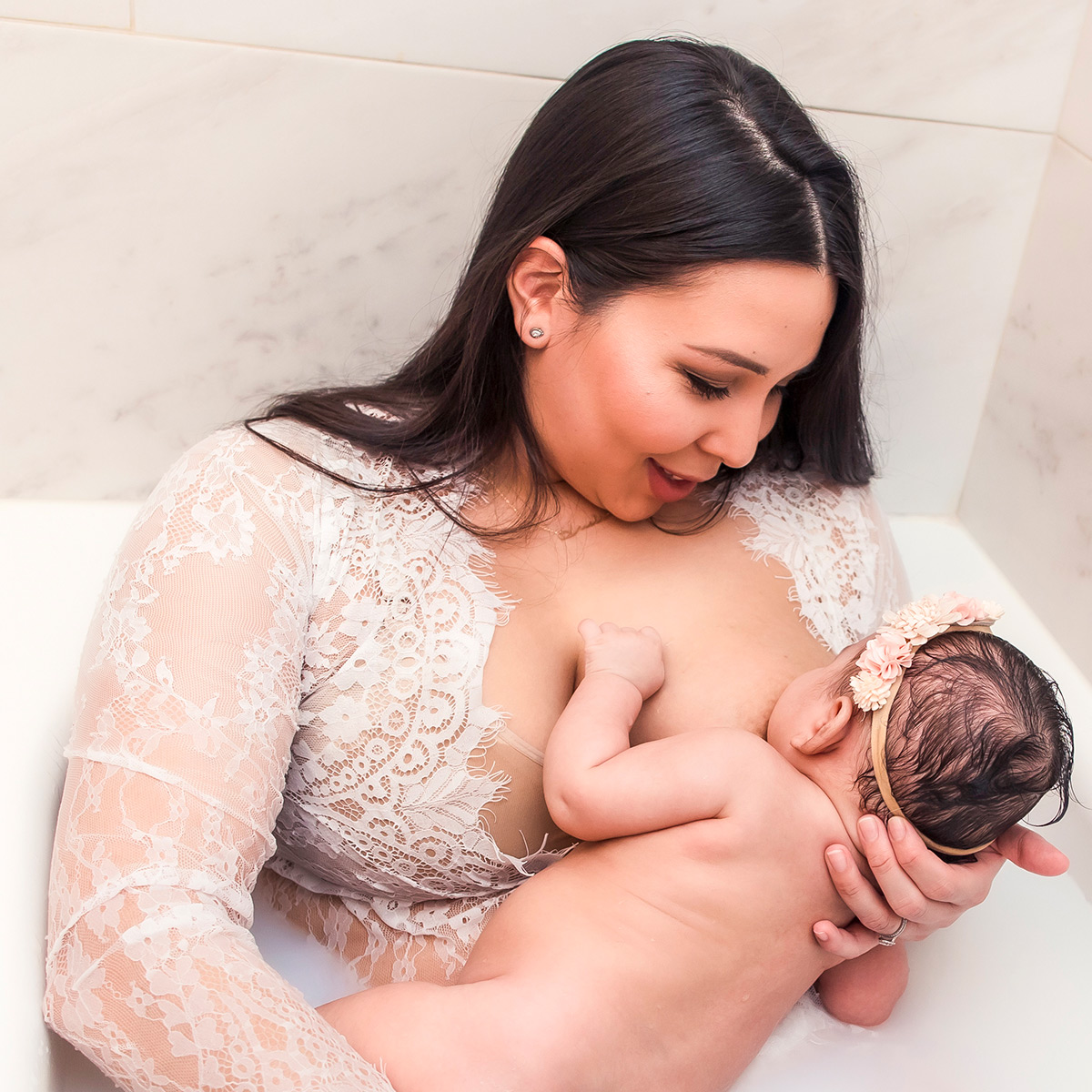 Woman in lace dress breastfeeing an infant in a bathtub.