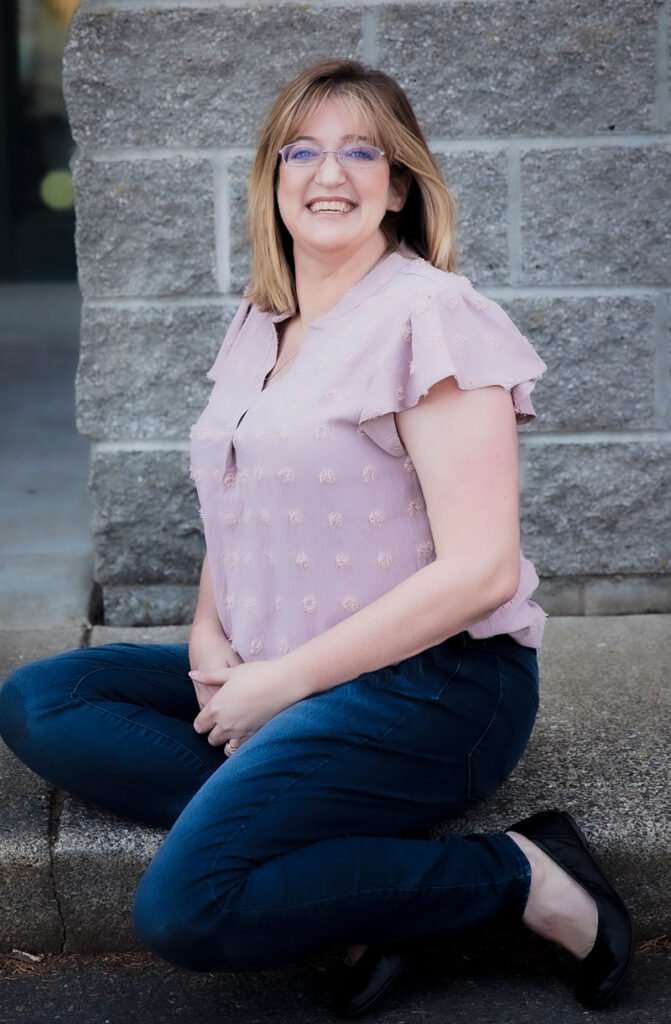 A woman with shoulder-length blonde hair and glasses is sitting on a stone ledge against a brick wall. She is wearing a light pink blouse, dark jeans, and black shoes. She smiles brightly while looking at the camera.